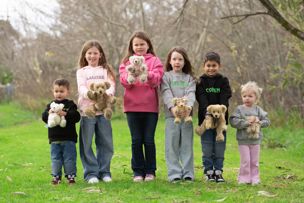 Six children standing outdoors on a grassy lawn with bare trees in the background, each holding a small teddy bear. The children are wearing casual clothing in pink, gray, black, and blue tones.